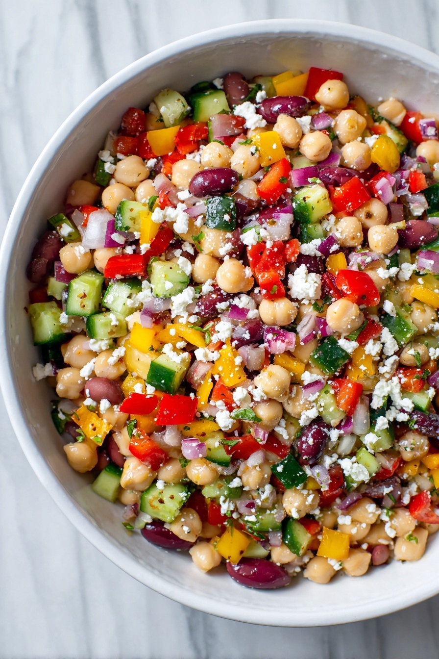 A white bowl filled with a colorful bean salad showing multiple layers of small, chopped vegetables and legumes. The base layer is made of light beige chickpeas and white beans mixed with a scattering of deep purple olive slices. On top, there is a thick mix of small diced bright red bell peppers, yellow bell peppers, green cucumbers, and red onions, creating a vibrant patchwork of colors. White crumbled cheese is sprinkled evenly over the salad, adding a soft, crumbly texture. The surface of the salad looks fresh, moist, and lightly coated in dressing. The bowl rests on a white marbled texture. Photo taken with an iphone --ar 2:3 --v 7 - Hearty Chickpea and Bean Salad, healthy bean salad, easy vegetarian salad, protein-packed salad recipes, mediterranean bean salad