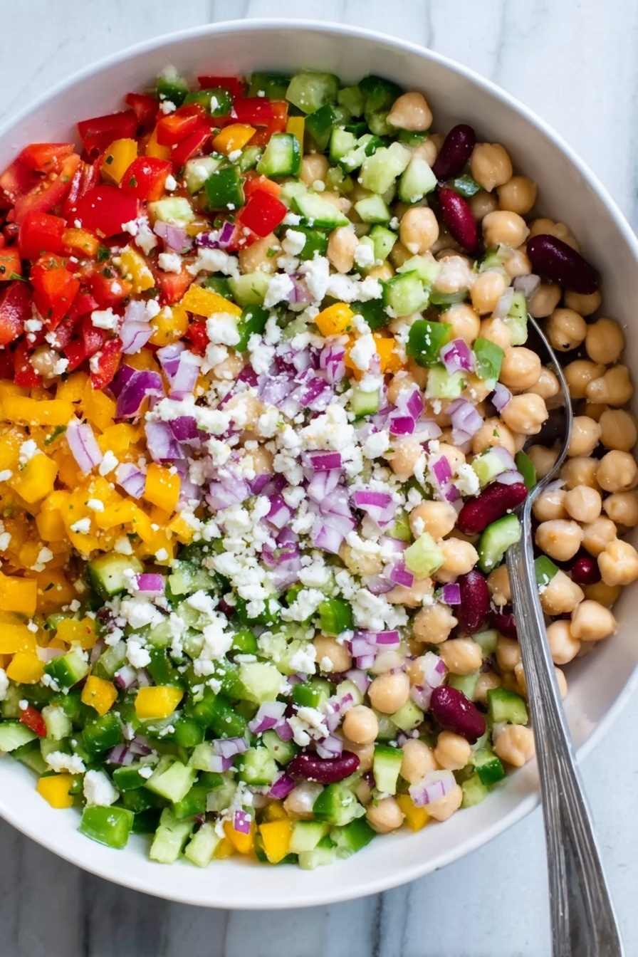The image shows a white bowl filled with a colorful salad made of many small chopped vegetables and beans. There are five visible layers: at the bottom is a mix of small round chickpeas and white beans with a creamy texture, above that are chopped bright red tomatoes and diced yellow peppers spread evenly, next is a layer of finely chopped green celery and cucumber giving a fresh look, on top of that are small pieces of purple onions scattered across, and sprinkled all over are tiny white crumbles of cheese. A silver spoon is resting on the left side inside the bowl. The bowl sits on a white marbled surface. Photo taken with an iphone --ar 2:3 --v 7 - Hearty Chickpea and Bean Salad, healthy bean salad, easy vegetarian salad, protein-packed salad recipes, mediterranean bean salad
