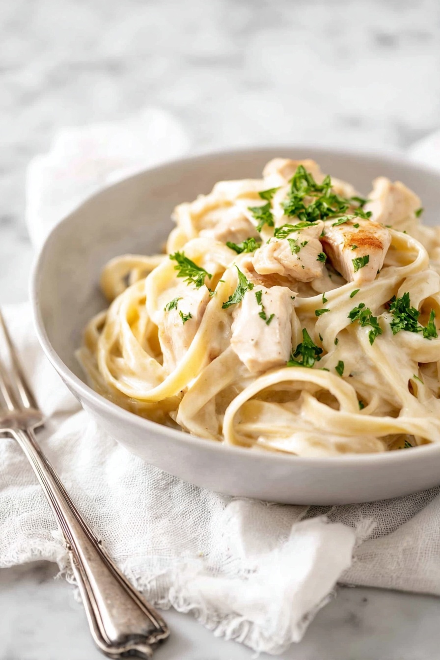 A white bowl holds creamy fettuccine pasta topped with pieces of light golden cooked chicken. The pasta is thick and pale, coated with a smooth white sauce. There are small green bits of parsley sprinkled on top, adding a fresh touch. The bowl sits on a soft white cloth, placed on a white marbled surface. Nearby, a silver fork rests on the cloth. photo taken with an iphone --ar 2:3 --v 7 - One-Pot Chicken Alfredo, Chicken Alfredo Pasta, Easy Creamy Alfredo, One-Pan Pasta Recipe, Simple Chicken Alfredo