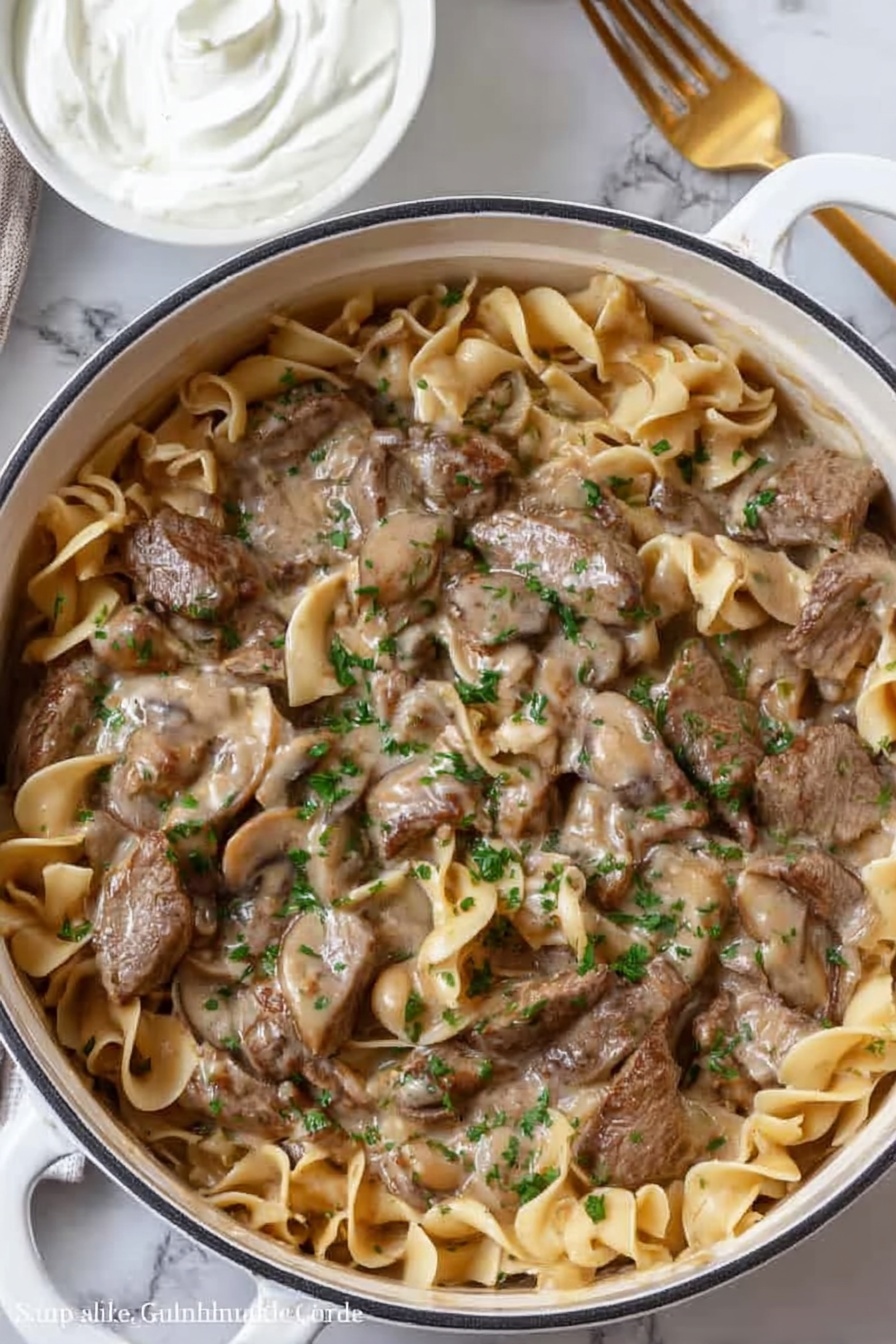 A white pot filled with a thick brown sauce that has visible pieces of orange carrots and sliced mushrooms within it. On top of the sauce, there is a pile of dark brown cooked strips of meat, arranged loosely in the center. The pot is placed on a white marbled surface. Nearby are two silver salt and pepper shakers and a white cloth with light gray stripes partially visible on the left side. photo taken with an iphone --ar 2:3 --v 7 - Easy Beef Stroganoff, beef stroganoff recipe, quick beef stroganoff, creamy mushroom beef, comfort food recipes