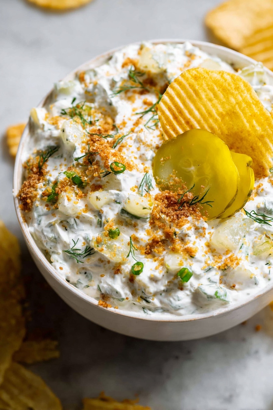 A white bowl filled with a creamy white dip mixed with small green herb pieces and light yellow chunks, topped with bright yellow pickle slices near the center. The dip is sprinkled with light brown crumbs and small green onion pieces, with a few small green herb sprigs scattered on top. A ridged golden potato chip is dipped into the creamy mixture near the edge of the bowl. The bowl sits on a white marbled surface with some crumbs and chips around it. photo taken with an iphone --ar 2:3 --v 7 - Ranch Pickle Dip, creamy pickle dip, party dip recipes, easy appetizer ideas, game day snack recipes