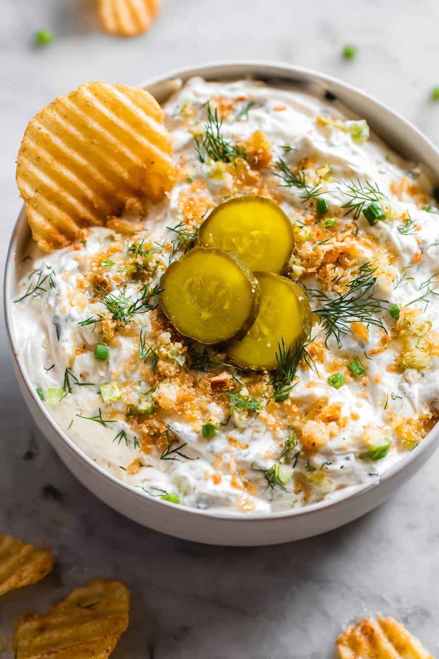 The image shows a grey bowl filled with a creamy white dip that is topped with several layers: small green chopped herbs, light brown crunchy bits, and three thick slices of pickles with a shiny, oily surface in the center. The dip looks thick and textured, with the crunchy bits scattered evenly on top. A woman’s hand is dipping a ridged, yellow chip into the creamy white mixture on one side. The bowl sits on a white marbled surface with some chips placed around it. photo taken with an iphone --ar 2:3 --v 7 - Ranch Pickle Dip, creamy pickle dip, party dip recipes, easy appetizer ideas, game day snack recipes