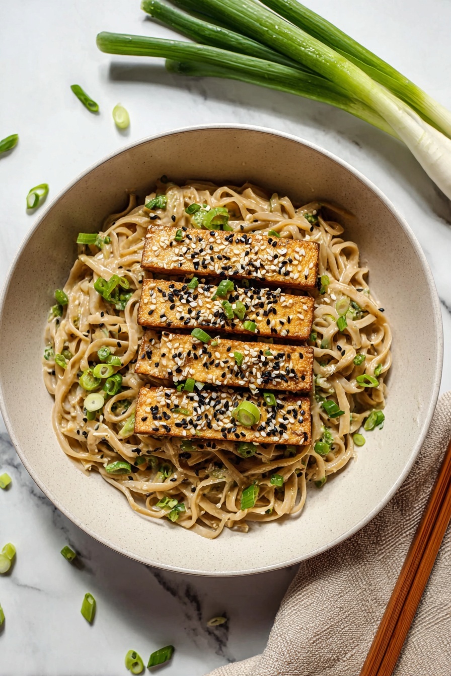 The image shows a close-up of a bowl with three layers. The bottom layer is thick, light tan noodles with a creamy sauce, speckled with tiny black and white seeds. On top of the noodles, there are golden-brown rectangular tofu pieces sprinkled with white and black sesame seeds and chopped green onions. At the top, a pair of wooden chopsticks held by a woman's hand lifts some noodles, with a few green onion slices stuck to the chopsticks. The bowl is white, and the background surface has a white marbled texture. photo taken with an iphone --ar 2:3 --v 7 - Peanut Udon Noodles, easy Asian noodle dish, quick vegetarian dinner, creamy peanut noodle recipe, flavorful udon stir-fry