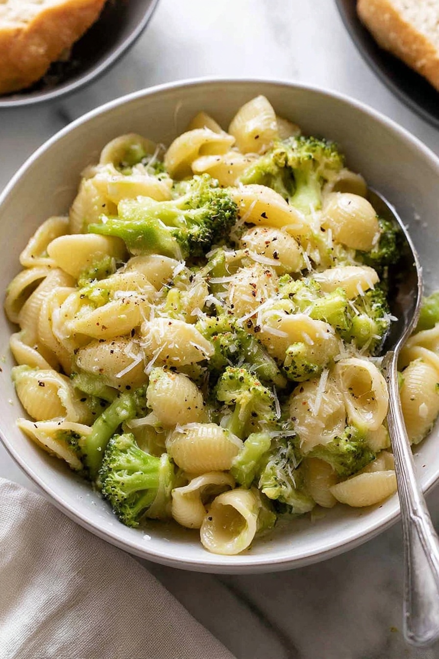 The image shows a white bowl filled with small shell pasta mixed with bright green broccoli pieces. The pasta looks soft and is coated with a light creamy sauce that makes the dish look smooth and shiny. There is a sprinkle of finely grated cheese on top and tiny specks of black pepper scattered over the dish. A silver spoon is placed partially inside the bowl on the right side. The bowl is set on a white marbled surface with a light cloth nearby and a blurred part of another dish with bread in the background. Photo taken with an iphone --ar 2:3 --v 7 - Quick Broccoli Pasta with Lemon and Cheese, healthy quick pasta recipes, easy broccoli pasta dinner, speedy veggie pasta, lemon cheese pasta dish