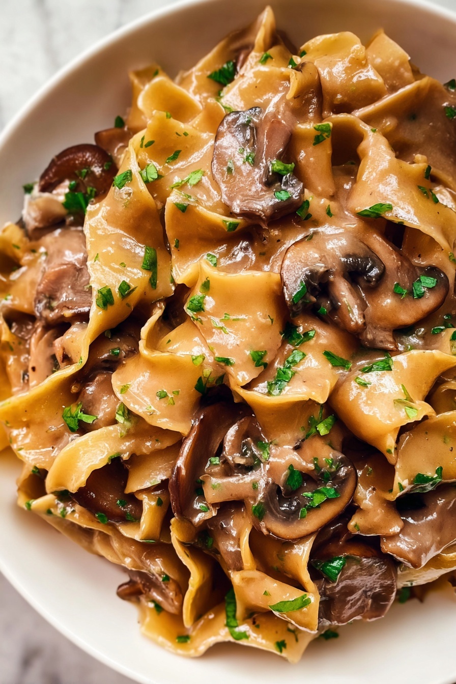 The image shows a close-up of a creamy pasta dish with wide, flat egg noodles mixed with thick slices of brown mushrooms. The noodles are coated in a smooth, light brown sauce that looks rich and glossy. Small pieces of chopped green herbs are sprinkled over the top, adding a fresh touch to the dish. The textures of the soft noodles and tender mushrooms blend together, showing a warm and comforting meal. The dish is served in a white bowl placed on a white marbled surface. photo taken with an iphone --ar 2:3 --v 7 - Vegetarian Mushroom Stroganoff, mushroom stroganoff recipe, easy vegetarian dinner, creamy mushroom sauce, quick weeknight vegetarian meal
