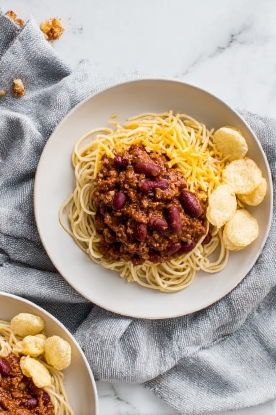 The image shows a white plate with three main layers. The bottom layer is a pile of light brown spaghetti noodles arranged in a loose small mound. On top of the noodles, there is a thick layer of dark reddish-brown chili with visible kidney beans and a chunky texture. To one side of the noodles and chili, there is a small group of round, pale beige crunchy chips neatly placed. The noodles and chips sit on a white marbled surface with a soft grey cloth draped around the plate. In the corner of the photo, part of another similar plate with the same food is visible. Photo taken with an iphone --ar 2:3 --v 7 - Cincinnati Chili Over Spaghetti, Cincinnati chili recipes, easy Cincinnati chili, Cincinnati-style chili, comfort food spaghetti dishes