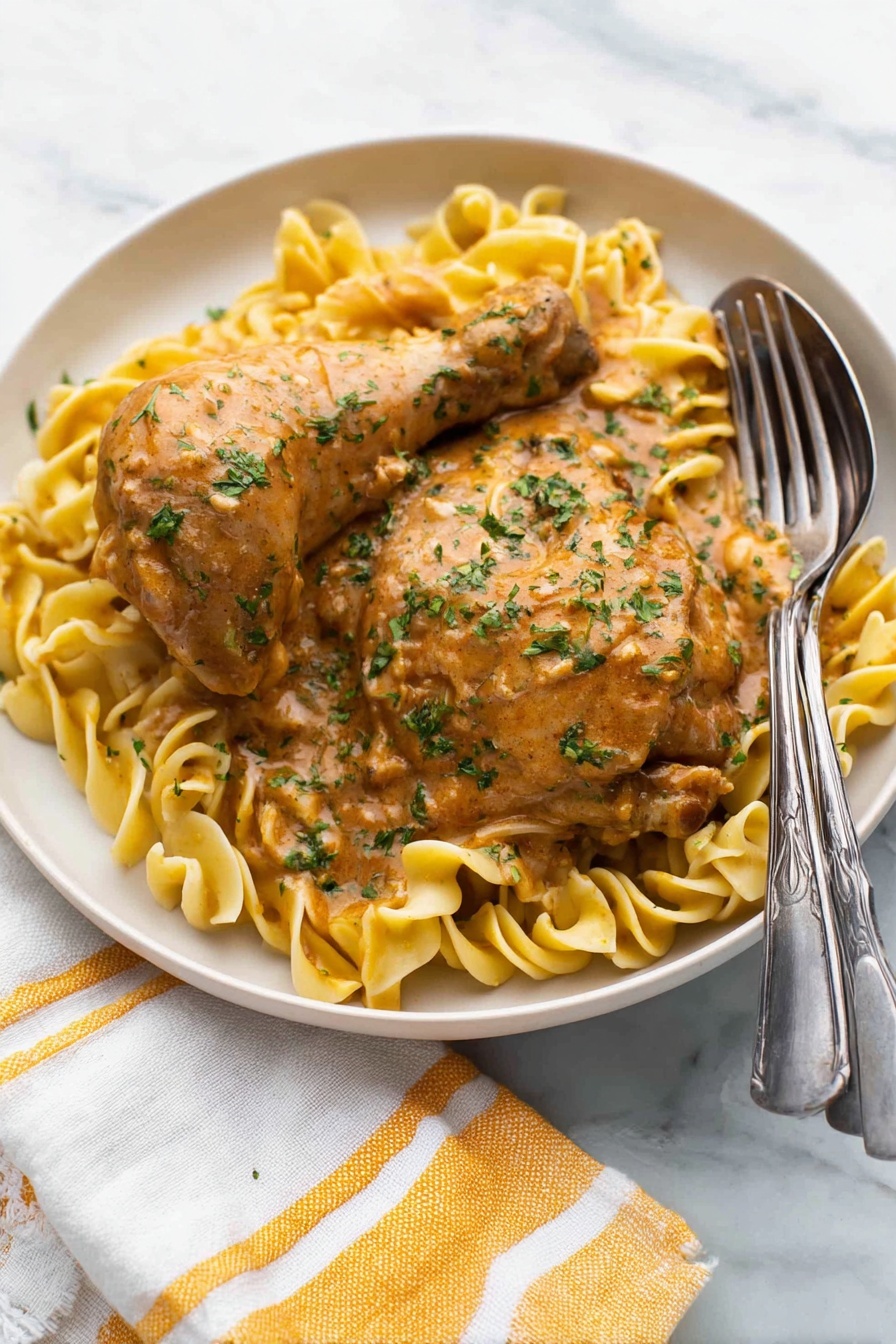 A white round plate holds two pieces of cooked chicken, one drumstick and one thigh, both covered in a thick light brown sauce with green herbs sprinkled on top. Underneath, there is a bed of light yellow egg noodles twisted softly, also coated lightly with the same sauce. A silver fork and spoon rest on the right edge of the plate. The plate sits on a white marbled surface with a white and yellow striped cloth napkin partially visible at the bottom left corner. Photo taken with an iphone --ar 2:3 --v 7 - Authentic Chicken Paprikash, Hungarian Chicken Paprikash, Chicken Paprikash recipe, traditional Hungarian dish, easy Chicken Paprikash