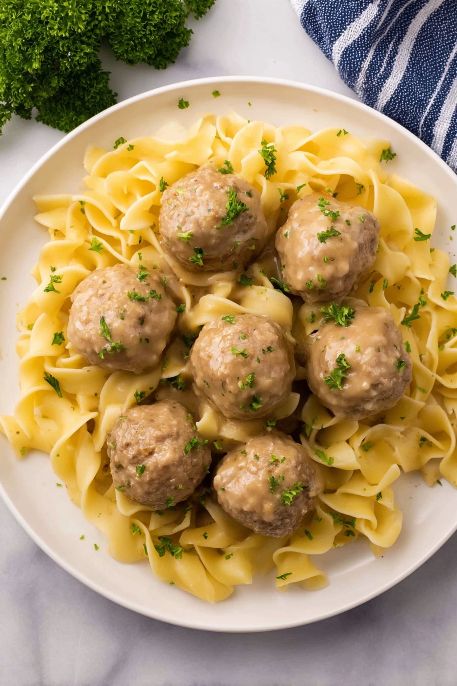 A white plate holds a dish with two main layers: six smooth, round beige meatballs evenly spread around the plate and a bed of soft, folded yellow egg noodles underneath and among the meatballs. The noodles have a slightly glossy texture and appear coated in light gravy. Both meatballs and noodles are lightly sprinkled with finely chopped green parsley, adding a fresh touch of color. The plate is set on a white marbled surface with a small bunch of parsley in the top left corner and a blue-striped towel in the bottom left corner. Photo taken with an iphone --ar 2:3 --v 7 - Creamy Swedish Meatball Noodles Skillet, Swedish meatballs with creamy sauce, easy comfort food recipes, one-pan dinner ideas, family-friendly skillet meals