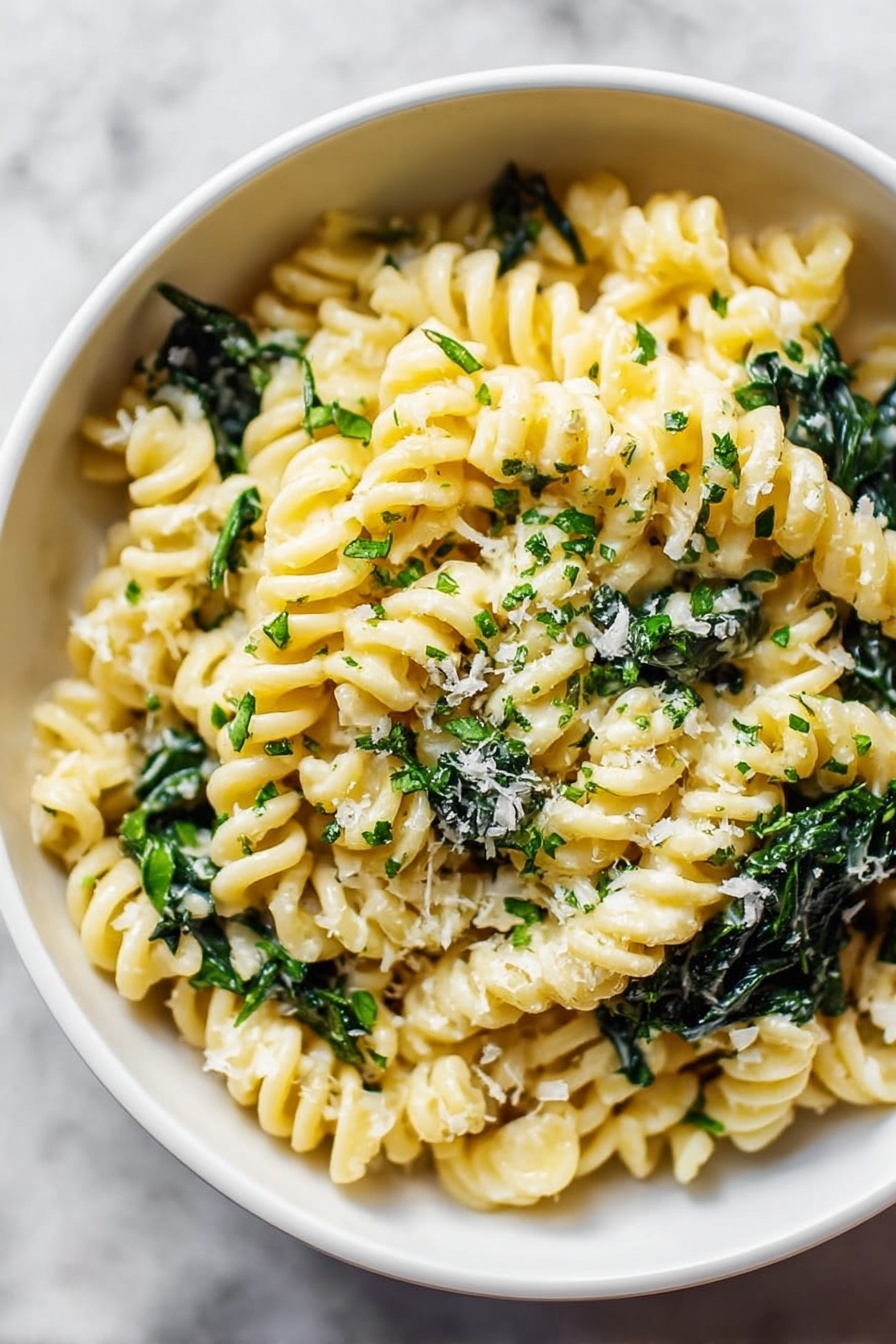 A close-up view of a bowl filled with short spiral pasta in a creamy light yellow sauce mixed with dark green leafy vegetables. The pasta is well coated and sprinkled lightly with small bits of grated white cheese and tiny chopped green herbs. The bowl is white and sits on a white marbled surface, and the overall look is fresh and simple. photo taken with an iphone --ar 2:3 --v 7 - Creamy Spinach Goat Cheese Pasta, spinach goat cheese pasta, creamy pasta with goat cheese, easy spinach goat cheese pasta, quick cheesy pasta with spinach
