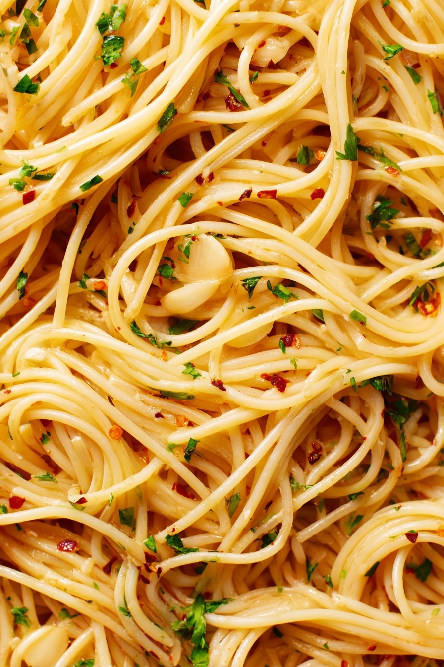 The image shows a close-up view of spaghetti pasta with thin, long noodles that are light golden in color. The noodles are mixed evenly with thin slices of light tan garlic, small bits of green parsley, and tiny red chili flakes scattered throughout. The spaghetti appears glossy with a slightly oily texture, making the noodles look smooth and shiny. The background is a white marbled texture. photo taken with an iphone --ar 2:3 --v 7 - Easy Spaghetti Aglio e Olio, Spaghetti Aglio e Olio, simple Italian pasta, quick garlic pasta, flavorful Italian spaghetti