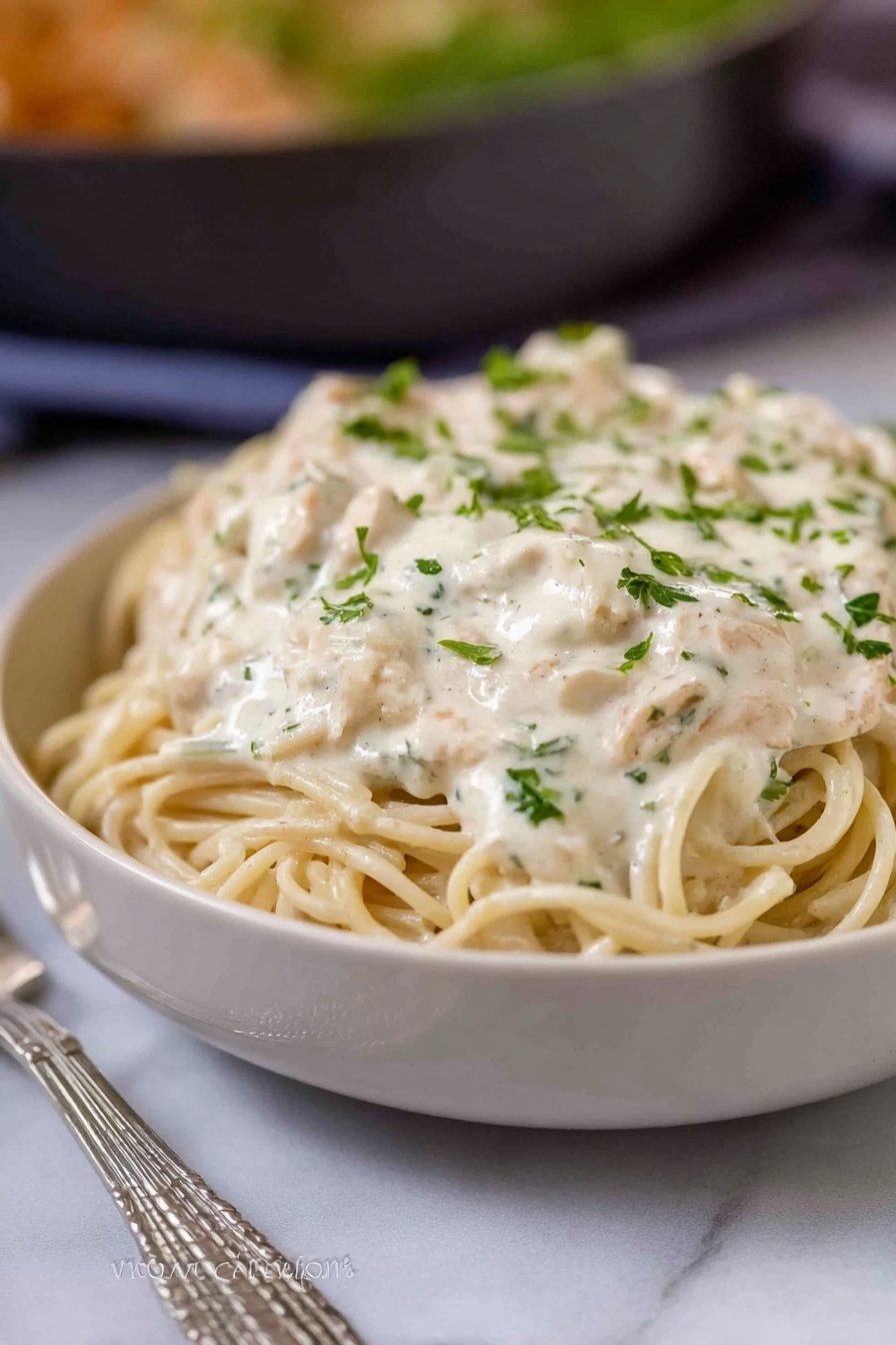 A close-up of a spoon lifting creamy pasta from a white bowl, showing three layers: the top layer is melted light golden cheese stretching with strings, the middle layer is creamy sauce covering small pieces of light beige chicken and thin slices of greyish mushrooms, all mixed with finely chopped bright green parsley, and the bottom layer is soft, pale yellow pasta strands coated in the sauce. The background is a white marbled texture, softly blurred to focus on the spoonful of food. Photo taken with an iphone --ar 2:3 --v 7 - Creamy Turkey Tetrazzini Pasta Bake, Turkey tetrazzini casserole, cheesy turkey pasta bake, hearty turkey pasta recipe, leftover turkey dinner ideas
