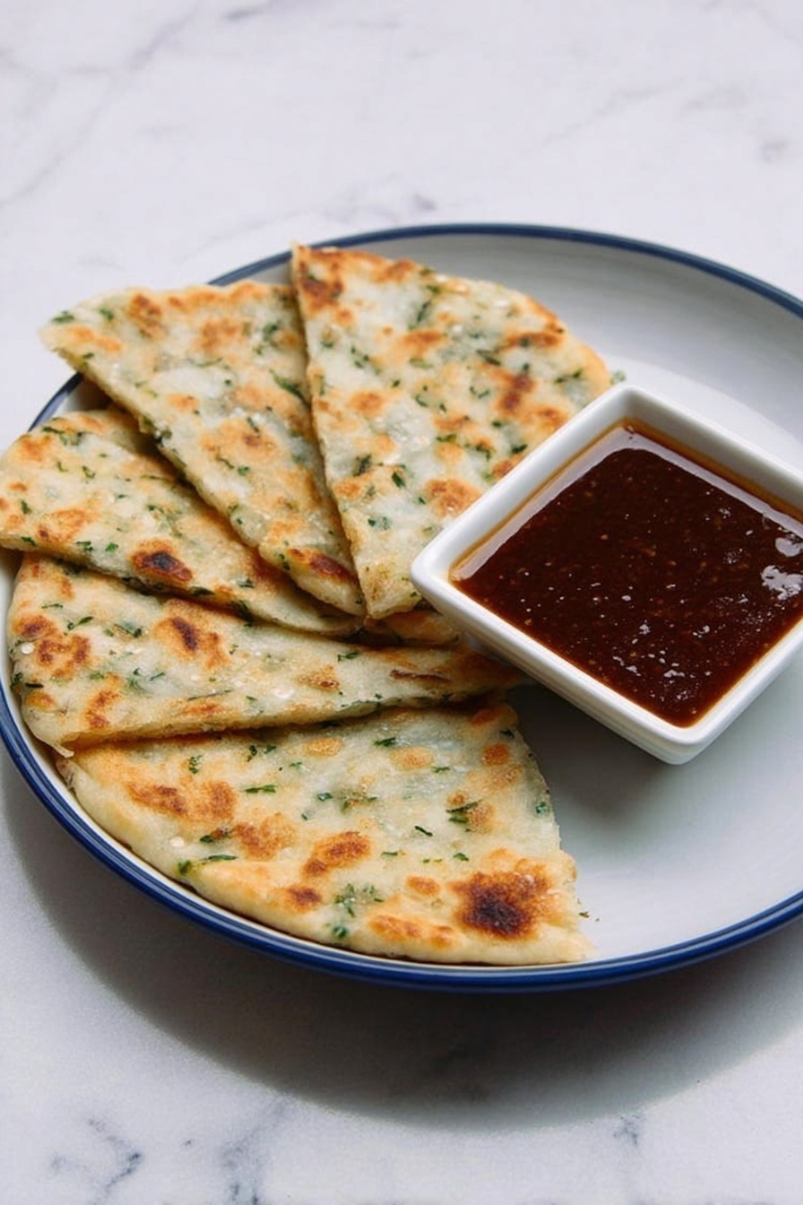 The image shows four pieces of flatbread-like food arranged in a fan shape on a white plate with a blue inside. The flatbreads are light golden-brown with some green herbs visible in the dough, showing a soft and slightly bumpy texture. On the plate, near the upper-right side of the flatbreads, there is a small white square bowl filled with a dark brown sauce that looks smooth and glossy. The plate is placed on a white marbled surface, creating a clean and simple background. photo taken with an iphone --ar 2:3 --v 7 - Chinese Scallion Pancakes, scallion pancake recipe, homemade scallion pancakes, crispy scallion pancakes, savory scallion pancakes