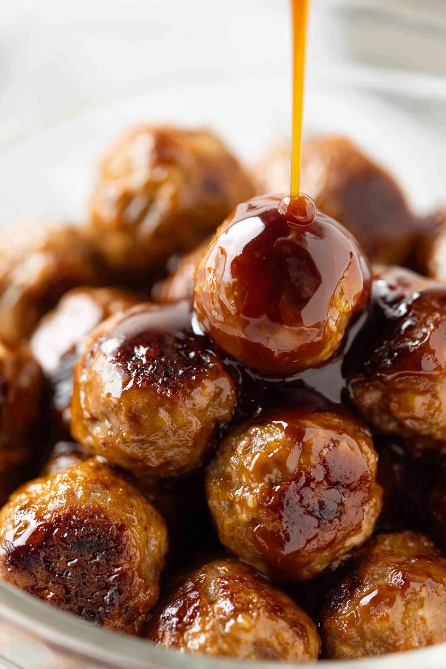 A close-up view of a pile of shiny brown meatballs in a clear bowl on a white marbled surface, with a thick reddish-brown sauce being poured slowly over the top meatball in the center. The meatballs are round and vary slightly in size, with a glazed texture showing small char marks and a glossy finish from the sauce. The sauce is thick and smooth, creating a shiny coating that drips down the sides of the meatballs. The image focuses closely on the meatballs with a soft blurry background. Photo taken with an iphone --ar 2:3 --v 7 - Baked Cocktail Meatballs with Sweet and Sour Sauce, party meatball recipes, easy appetizer ideas, homemade sweet and sour sauce, juicy meatball recipes