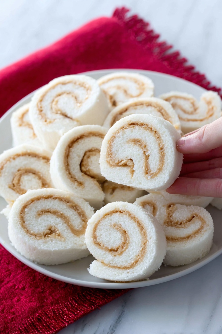 The image shows a white plate filled with soft, spiral-shaped rolls arranged closely together. Each roll has a smooth white outer layer with swirled light tan filling inside, creating a clear contrast. The rolls look fluffy and neatly sliced, with the spirals visible on the sides. The plate is placed on a bright red cloth, and one roll is in the foreground, slightly separated. The background and surface have a white marbled texture. A woman's hand is holding one roll in the top right corner. photo taken with an iphone --ar 2:3 --v 7 - Potato Candy Rolls with Peanut Butter, nostalgic candy recipes, no-bake potato candy, homemade peanut butter treats, old-fashioned candy ideas