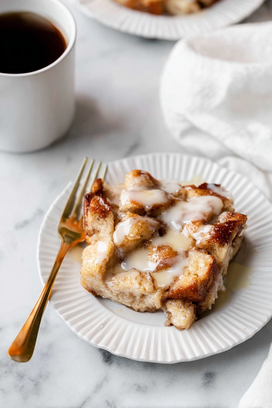 A piece of bread pudding with chunky, soft golden pieces of baked bread layered unevenly, soaked in a creamy, slightly shiny white sauce drizzled on top. The bread looks light brown with some darker toasted edges, sitting on a white plate with a subtle scalloped edge. A gold fork is placed on the left side of the plate. The plate rests on a white marbled surface, and a soft white cloth is partially visible in the upper right corner. There is also a white cup filled with dark coffee in the top left corner. photo taken with an iphone --ar 2:3 --v 7 - Apple Pie Cinnamon Roll Bake, cinnamon roll breakfast bake, apple pie cinnamon roll, easy apple cinnamon breakfast, fall brunch recipes