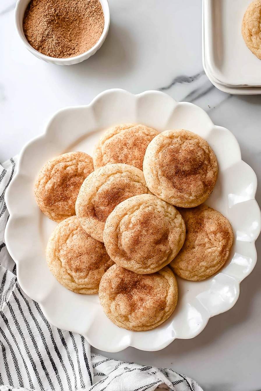 There are seven round snickerdoodle cookies with a light golden brown color and a dusting of cinnamon sugar on top, arranged in a flower shape on a white scalloped plate. The cookies have a soft, slightly wrinkled texture and are placed on a white marbled surface. Near the top left of the plate, there is a small white bowl filled with cinnamon sugar. A white cloth with thin black stripes lays slightly under the plate on the left side. Photo taken with an iphone --ar 2:3 --v 7 - Chewy Snickerdoodle Cookies, cinnamon sugar cookies, soft chewy cookie recipe, homemade snickerdoodles, classic cinnamon cookies