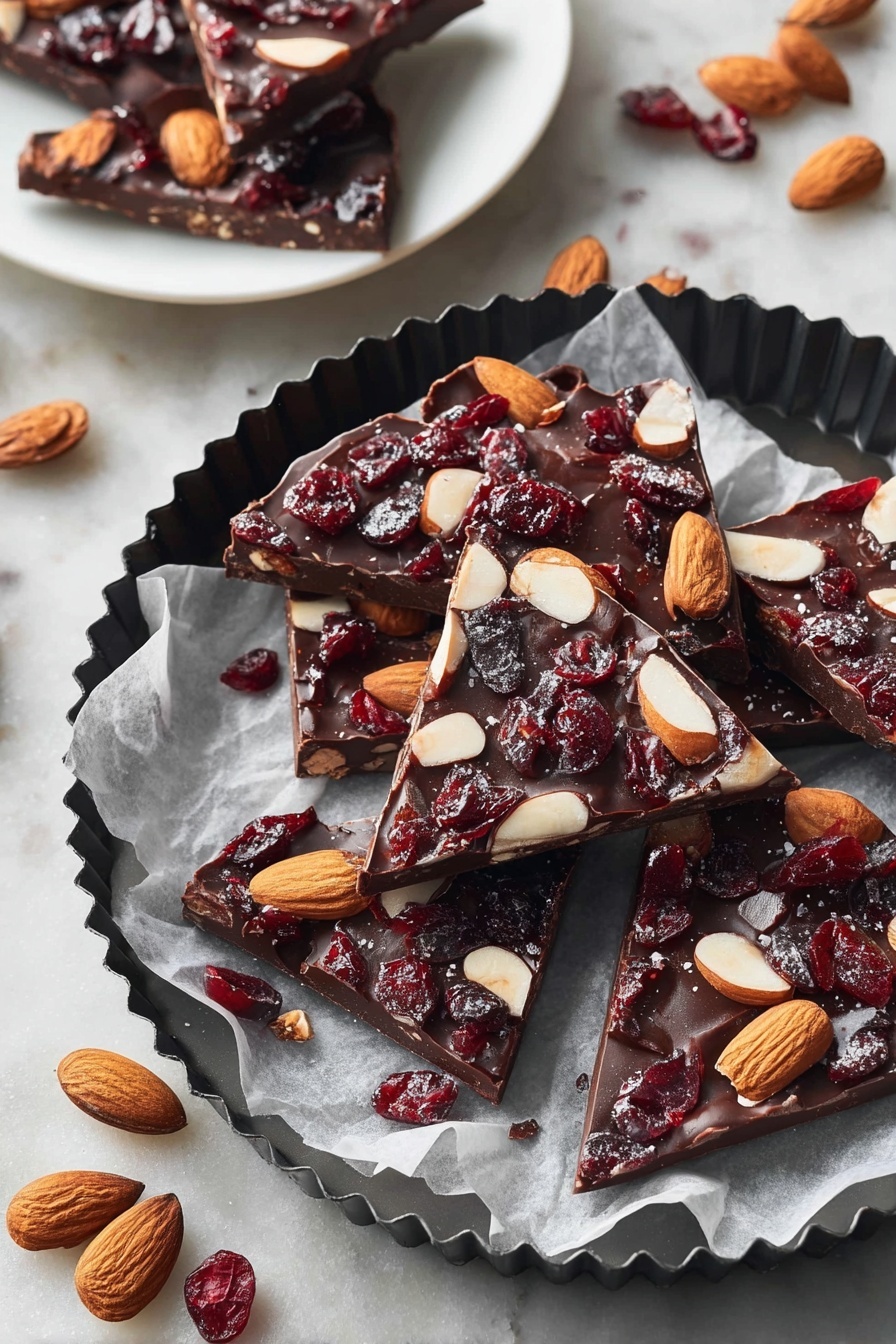 The image shows several pieces of chocolate bark, each with one thick layer of dark brown chocolate topped with whole light brown almonds and dark red dried cranberries scattered evenly. The chocolate pieces are irregular triangles and squares placed on white parchment paper inside a black fluted round pan. Around the pan are whole almonds and dried cranberries on a white marbled textured surface. In the background, a white plate holds three more pieces of chocolate bark, showing the same dark chocolate base with almonds and cranberries. photo taken with an iphone --ar 2:3 --v 7 - Easy Chocolate Bark, Chocolate Bark with Nuts and Fruit, Nut and Fruit Chocolate Bark, Quick Chocolate Bark, Homemade Chocolate Treats