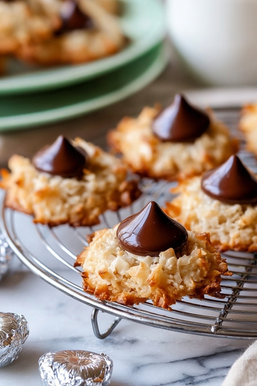 The image shows several round cookies with a rough, golden-brown texture from toasted coconut flakes, each with a smooth, shiny dark chocolate drop placed in the center. The cookies sit on a silver wire cooling rack, which rests on a white marbled surface. Around the rack, there are small foil-wrapped chocolate drops scattered. The background includes a blurred white bowl and a soft green plate with more cookies, creating a warm and inviting scene. photo taken with an iphone --ar 2:3 --v 7 - Coconut Macaroon Blossoms with Chocolate Kisses, coconut macaroon cookies, chocolate kiss cookies, gluten free coconut cookies, easy holiday cookie recipes