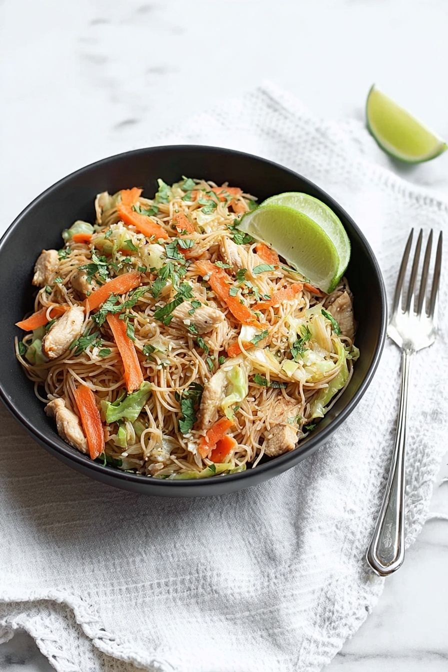 A black bowl sits on a white marbled surface with a white cloth beneath it, filled with thin light brown noodles mixed with small pieces of light beige chicken, bright orange carrot sticks, light green cabbage, and some fresh green herbs scattered on top. A half lime with a pale green inside rests on the right edge of the bowl. To the right of the bowl are a silver spoon and fork placed on the white marbled surface. photo taken with an iphone --ar 2:3 --v 7 - Easy Chicken and Pork Pancit, Filipino Pancit Recipe, Simple Chicken and Pork Noodles, Quick Filipino Stir-Fry, Savory Pancit with Chicken and Pork