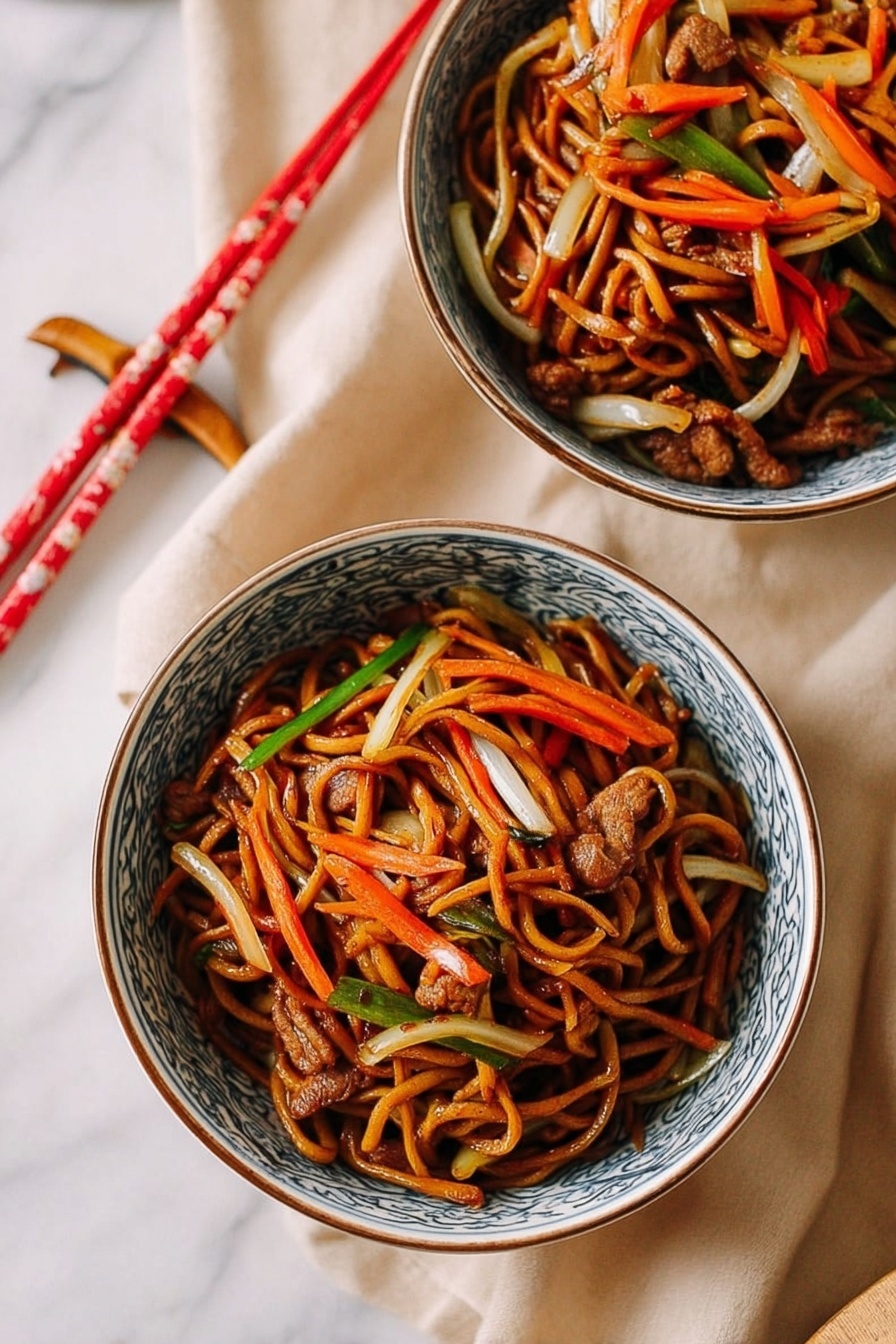 Two white bowls with blue patterns on the edges hold a serving of stir-fried noodles. The dish has multiple layers: a base of dark brown soy-coated noodles entwined with thin strips of orange carrots, white onions, and light green vegetables. Small pieces of cooked brown meat are scattered throughout. Both bowls are placed on a soft beige cloth on a white marbled surface. A pair of red and cream chopsticks rest near the bowls. photo taken with an iphone --ar 2:3 --v 7 - Restaurant-Style Chicken Lo Mein, chicken lo mein recipe, Chinese chicken noodle stir-fry, homemade lo mein with chicken, easy Chinese chicken lo mein