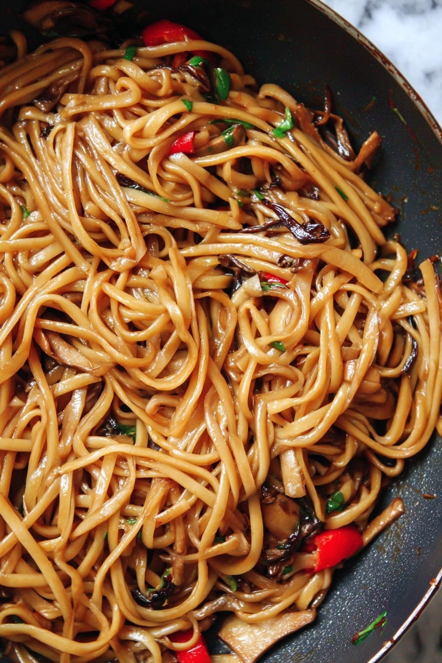 The image shows a close-up of a pan filled with long, shiny noodles coated in a light brown sauce. Mixed among the noodles are small pieces of light tan tofu, dark brown thin mushroom strips, and bright red chili slices, with tiny bits of green herbs scattered throughout. The noodles look soft and glossy, with a slight twist and curl to them, giving a textured, tangled appearance. The pan has a dark surface and the background is changed to a white marbled texture. photo taken with an iphone --ar 2:3 --v 7 - Vegan Kung Pao Pasta with Mushrooms, vegan pasta recipes, vegan Asian fusion dishes, plant-based pasta with mushrooms, spicy vegan pasta