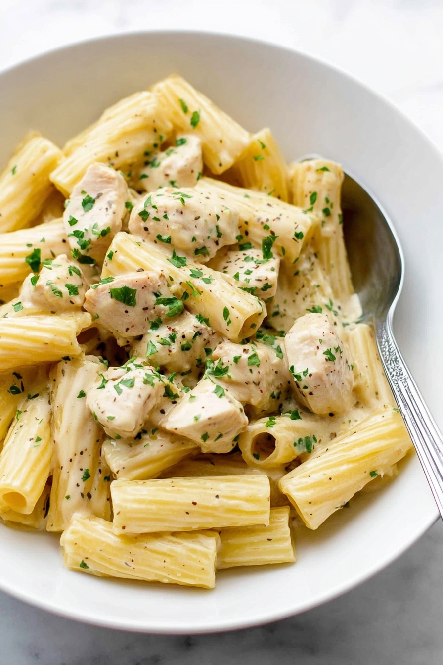 A white bowl filled with creamy rigatoni pasta mixed with chunks of light beige chicken pieces, all coated in a smooth, pale yellow sauce. The pasta is ribbed and tubular, scattered evenly throughout the dish. Small green parsley flakes are sprinkled over the top, adding bright color contrast. A silver spoon rests inside the bowl on the right side, partially under the pasta. The bowl sits on a white marbled surface. Photo taken with an iphone --ar 2:3 --v 7 - Creamy Lemon Chicken Pasta, Lemon Chicken Pasta, Easy Chicken Pasta Recipes, Quick Creamy Pasta, Lemon Chicken Dinner