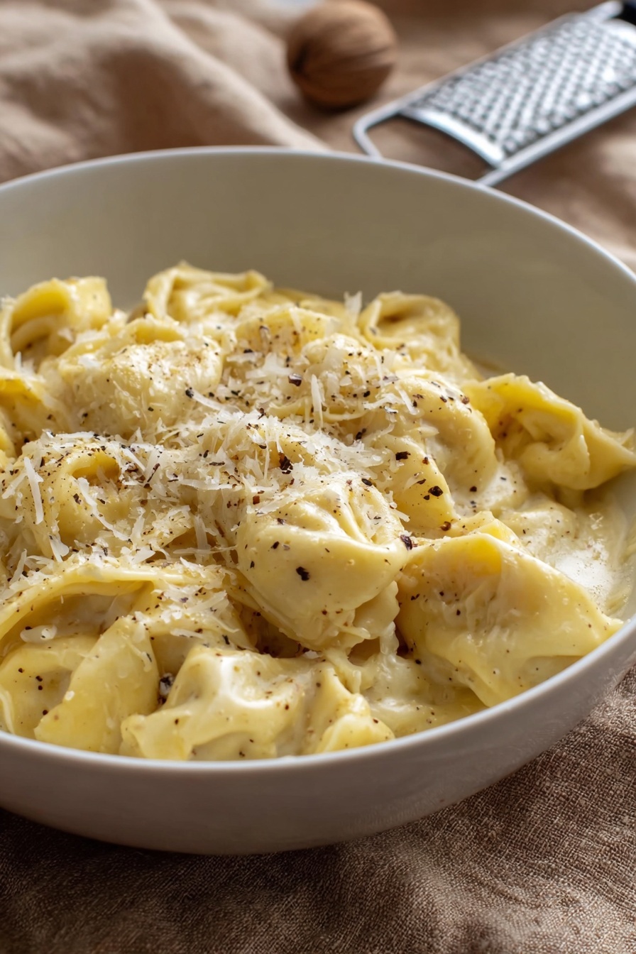 A white bowl filled with about two layers of soft, folded tortellini pasta in a creamy, pale yellow sauce. The pasta is sprinkled with finely grated white cheese and small black pepper flakes, mostly concentrated on the top center. The bowl rests on a tan textured cloth, with a metal grater and a whole nutmeg blurred in the background. The lighting is natural and soft, showing the smooth creamy texture of the sauce and the delicate folds of the tortellini photo taken with an iphone --ar 2:3 --v 7 - Creamy Cheese Tortellini, cheesy tortellini pasta, quick tortellini dinner, easy cheese tortellini recipe, indulgent pasta dishes