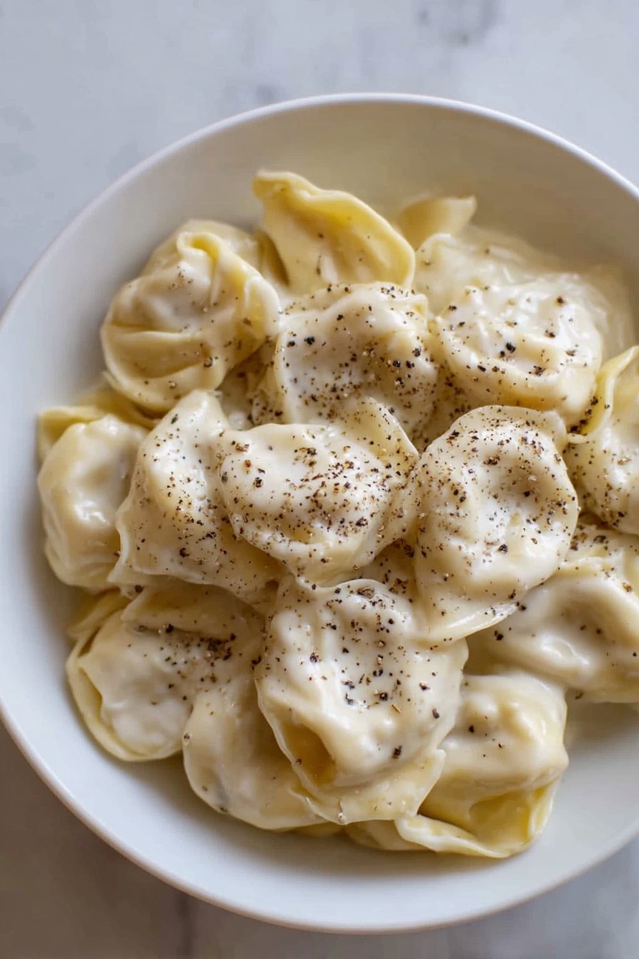 A white bowl filled with a single layer of tortellini pasta, covered evenly with a smooth, creamy white sauce that has a slightly shiny texture. The tortellini are pale yellow with soft folds, and black pepper is sprinkled generously on top, adding small dark specks scattered over the sauce. The bowl is placed on a white marbled surface, and the light highlights the soft texture of both the pasta and sauce photo taken with an iphone --ar 2:3 --v 7 - Creamy Cheese Tortellini, cheesy tortellini pasta, quick tortellini dinner, easy cheese tortellini recipe, indulgent pasta dishes