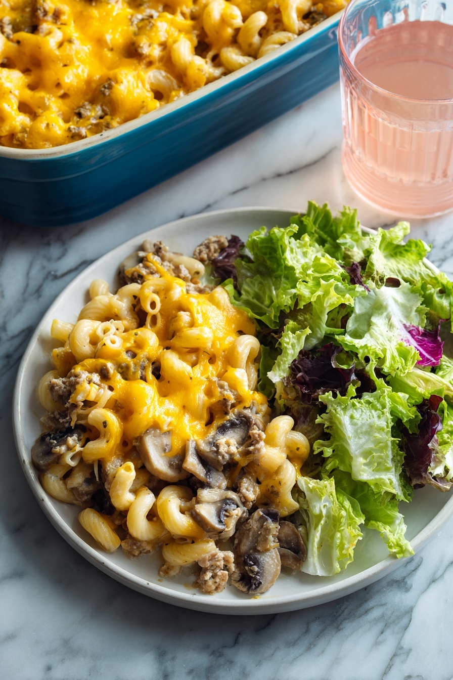 The image shows a white round plate with two main parts: on the left, a layer of baked macaroni mixed with ground meat and sliced mushrooms, all topped with melted bright yellow-orange cheese that looks gooey and soft, and on the right, a fresh green salad made of different lettuce leaves with a mix of light and dark green colors and some purple edges. The plate is set on a white marbled surface, and in the background, there is a blue baking dish filled with more of the macaroni bake and a clear glass of light pink drink. Photo taken with an iphone --ar 2:3 --v 7 - Chuck Roast Recipe, hearty beef dinner, slow cooker chuck roast, tender beef recipes, comfort food ideas
