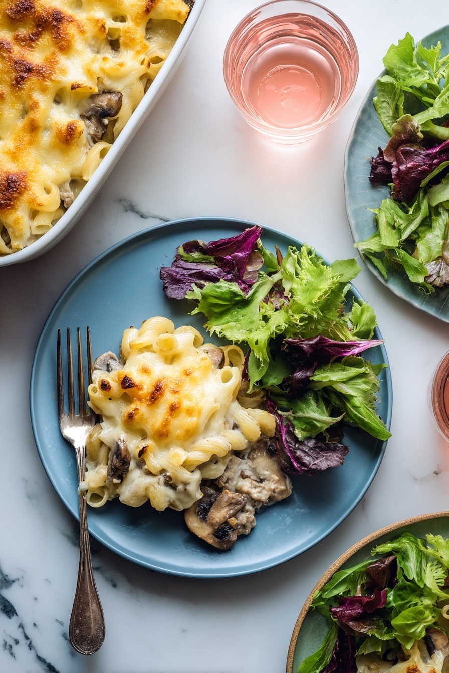 A blue plate holds a serving of baked macaroni and cheese with visible pieces of mushrooms and ground meat under a layer of melted golden-yellow cheese on the left side; on the right side of the plate, there is a fresh green salad made of mixed leafy greens with different shades of green and purple. A silver fork rests on the plate's left edge underneath the macaroni. In the background, a white baking dish filled with more macaroni and cheese sits on a white marbled surface along with a small clear glass of light pink drink. Another plate with leafy greens and another glass of pink drink are partially visible on the right side. photo taken with an iphone --ar 2:3 --v 7 - Chuck Roast Recipe, hearty beef dinner, slow cooker chuck roast, tender beef recipes, comfort food ideas