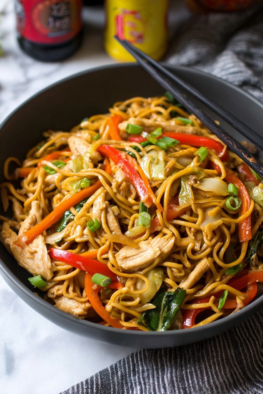 A close-up view of a dark gray bowl filled with stir-fried noodles mixed with thin strips of light beige chicken pieces, bright red bell pepper slices, green scallion pieces, thin orange carrot strips, light green cabbage, and sliced beige mushrooms. Black chopsticks rest on the right edge of the bowl, while the bowl sits on a white marbled surface with part of a gray striped cloth to the right. The background includes some blurred bottles with yellow and red labels. photo taken with an iphone --ar 2:3 --v 7 - Chicken Yakisoba Stir-Fry, Chicken Yakisoba, stir-fry dinner, Japanese-style noodle recipe, easy Japanese stir-fry