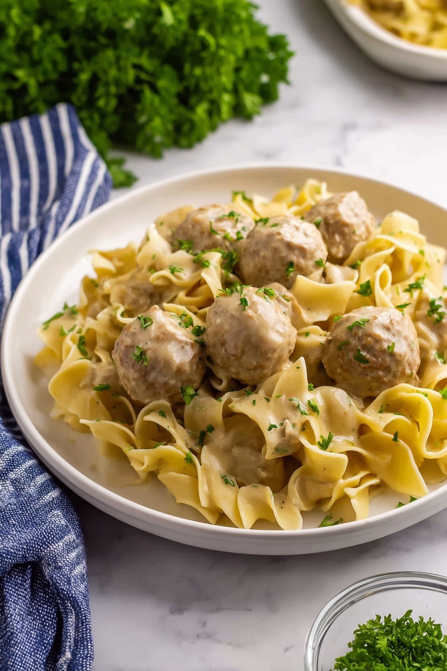 A white plate holds a serving of wide, curly egg noodles mixed with round meatballs covered in a creamy light brown sauce. The noodles are soft and slightly twisted, with flecks of green herbs sprinkled on top. The meatballs are light brown, smooth, and sit evenly spaced among the noodles. Bright green parsley leaves are scattered across the dish, adding color contrast. The scene is set on a white marbled surface with a bunch of fresh parsley blurred in the background, a blue and white striped cloth partially visible at the bottom left, and a small glass bowl with chopped herbs near the plate. Photo taken with an iphone --ar 2:3 --v 7 - Creamy Swedish Meatball Noodles Skillet, Swedish meatballs with creamy sauce, easy comfort food recipes, one-pan dinner ideas, family-friendly skillet meals