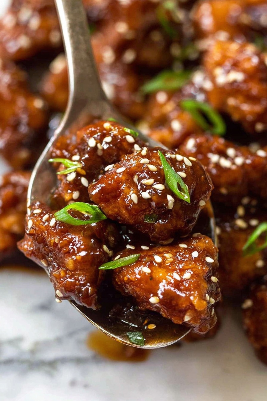 The image shows a close-up view of small pieces of fried chicken coated in a thick, shiny, dark brown sauce. The chicken pieces are irregular in shape with a crispy texture underneath the sauce. White sesame seeds are scattered on top, adding contrast and texture. Thin green slices of scallions or green onions are sprinkled over the chicken, providing a splash of fresh color. The chicken rests in a metal spoon, with more saucy chicken pieces visible in the background, all set on a white marbled surface. photo taken with an iphone --ar 2:3 --v 7 - Crispy General Tso's Chicken, General Tso's Chicken recipe, homemade Chinese chicken, Asian chicken stir-fry, crispy chicken dinner