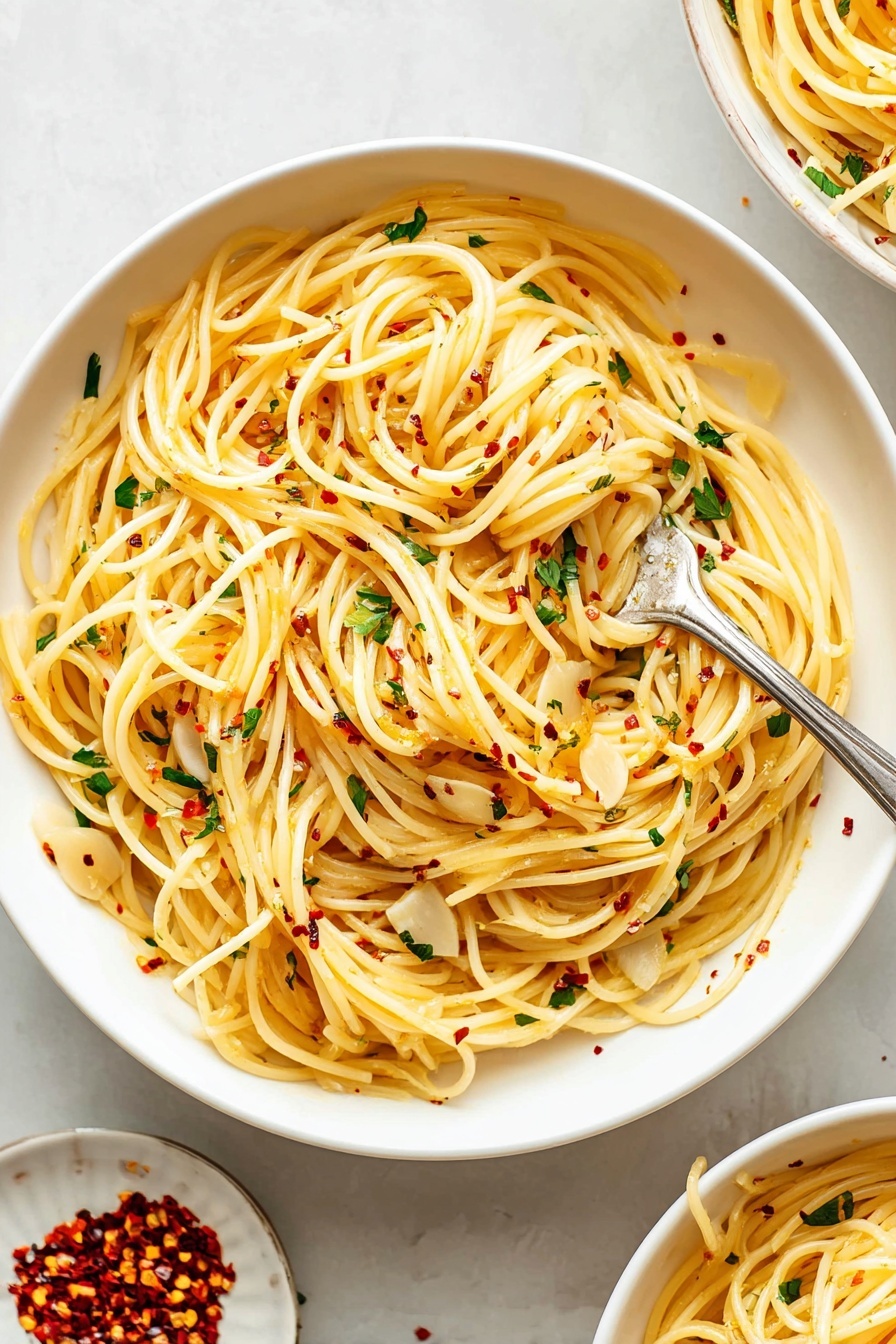 A large white bowl filled with long, thin spaghetti noodles that are lightly coated with olive oil, giving them a soft yellow color. Scattered throughout the pasta are thin, flat slices of garlic and small bits of fresh green parsley. Tiny red chili flakes are sprinkled evenly on top, adding small pops of red. A silver fork is twirling a small bundle of the pasta near the center of the bowl. The bowl rests on a white marbled surface, with a small white plate next to it containing additional red chili flakes. Part of another similar bowl filled with the same pasta can be seen in the upper right corner. Photo taken with an iphone --ar 2:3 --v 7 - Easy Spaghetti Aglio e Olio, Spaghetti Aglio e Olio, simple Italian pasta, quick garlic pasta, flavorful Italian spaghetti