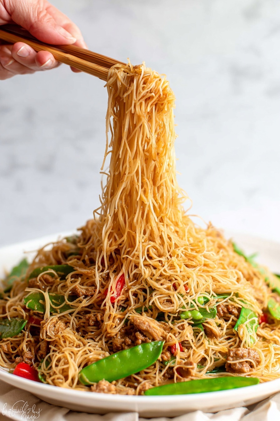 The image shows a white plate full of thin noodles mixed with small pieces of cooked meat, green snow peas, and red bell pepper slices. The noodles are light brown and soft, forming the top and main layer, while the vegetables and meat are scattered throughout, adding pops of green and red colors. A woman's hand is holding wooden chopsticks lifting a portion of the noodles above the plate, with the noodles hanging down in thin strands. The background has a white marbled texture. photo taken with an iphone --ar 2:3 --v 7 - Singapore Curry Noodles with Chicken, Singapore Curry Noodles, Curry Noodles with Chicken, Easy Singapore Curry Noodles, Quick Asian Noodle Recipes