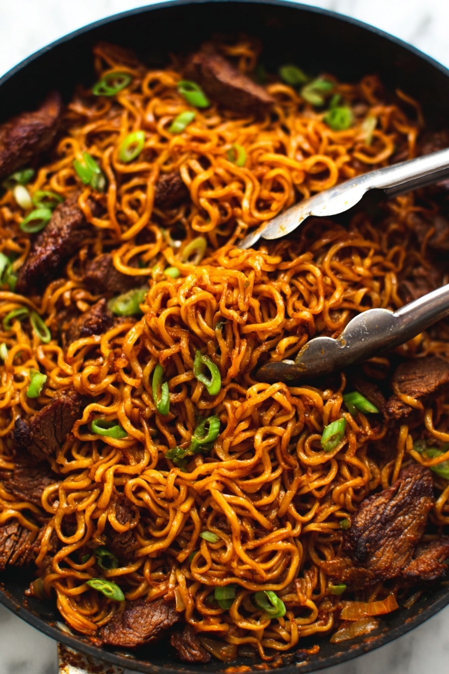 The image shows a close-up of a black pan filled with cooked noodles mixed with pieces of dark brown meat. The noodles are a warm orange-brown color, twisted and tangled together with small green onions sprinkled on top for color contrast. A pair of silver tongs is gripping some noodles and meat in the top right area of the pan. The pan sits on a white marbled surface. photo taken with an iphone --ar 2:3 --v 7 - Spicy Korean Beef Noodles, Korean Beef Noodles, Spicy Noodle Recipe, Korean Beef Stir-Fry, Quick Korean Noodles