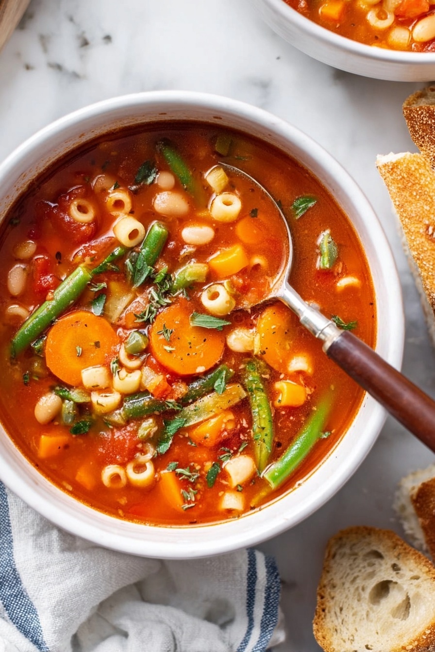 A white bowl filled with a bright red tomato-based soup with visible small round pasta pieces, sliced orange carrots, white beans, green beans, and finely chopped herbs. A silver spoon with a brown handle is placed inside the bowl, resting on the side. The bowl sits on a white marbled surface. To the right, rustic pieces of sliced bread are partially visible, and on the left, a soft white cloth with blue stripes is partly in the frame. photo taken with an iphone --ar 2:3 --v 7 - Vegetarian Minestrone Soup, healthy vegetarian soup, hearty vegetable soup, wholesome minestrone recipe, easy vegetarian soup