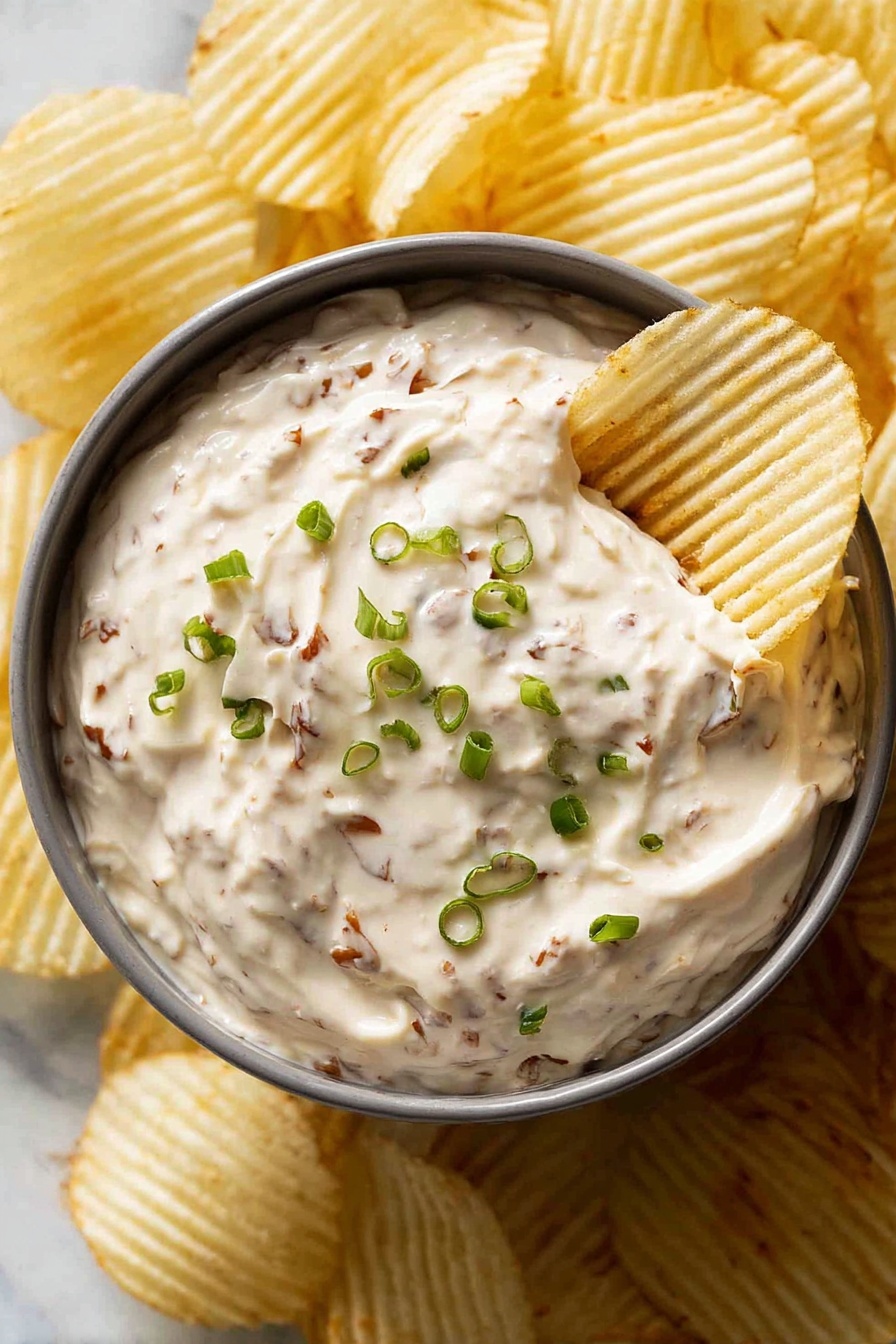 A gray round bowl filled with creamy dip that has visible small brown bits throughout, topped with finely chopped green onions scattered on the surface. A white ridged potato chip is dipped into the creamy mixture on the right side of the bowl. The bowl is surrounded by more white ridged potato chips, all arranged on a white marbled surface. photo taken with an iphone --ar 2:3 --v 7 - Homemade French Onion Dip, French Onion Dip, caramelized onion dip, creamy onion dip, party dip recipes