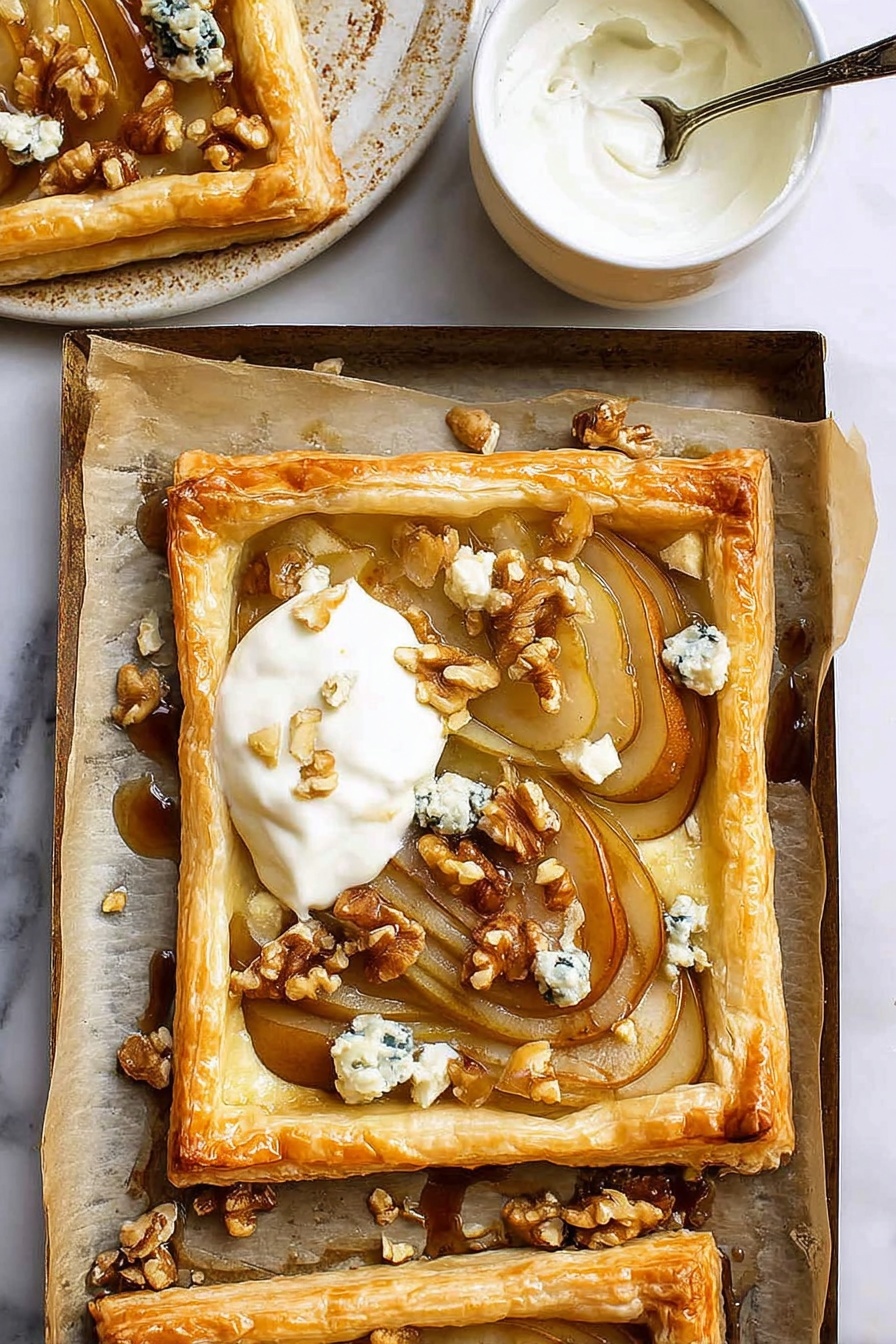 The image shows a golden brown puff pastry tart with a raised edge surrounding soft, thinly sliced caramelized pears arranged in an overlapping pattern. On top of the pears, there are small chunks of blue cheese and scattered walnut pieces adding texture and color contrast. A dollop of creamy white yogurt or cream is placed on one corner of the tart. The tart sits on parchment paper which lines a baking tray with some walnut pieces and cheese crumbs around it. Nearby, a small white ceramic bowl with a spoon inside contains more white cream. The background is a white marbled surface. photo taken with an iphone --ar 2:3 --v 7 - Caramelized Pear and Blue Cheese Tarts, pear and blue cheese tart, savory pear tart, blue cheese puff pastry tart, elegant fruit and cheese tart