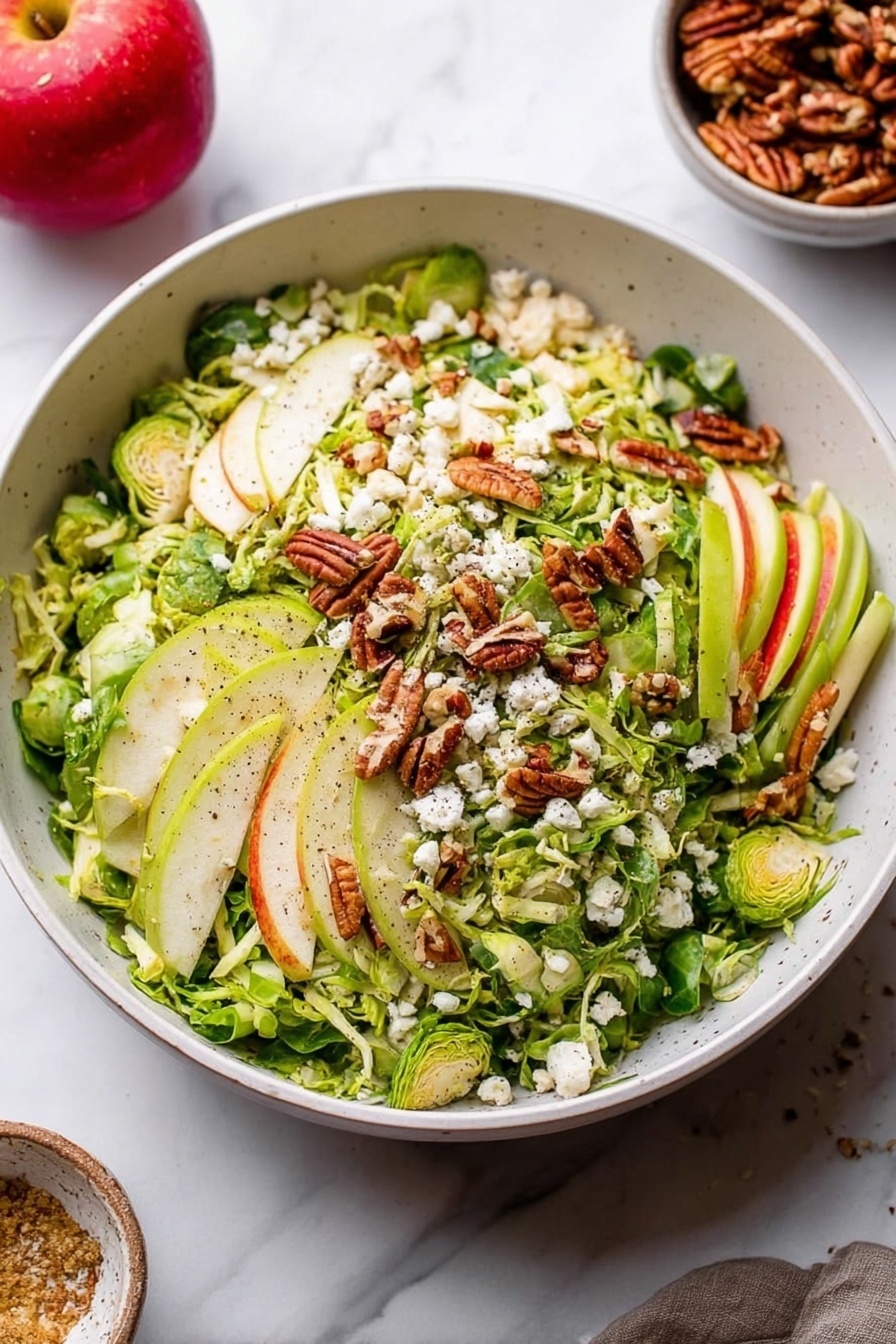 A large white bowl filled with a fresh salad featuring several layers: the base layer is thinly sliced green Brussels sprouts, topped with light green and white strips of apple. Scattered on top are golden-brown pecans and small white crumbles of cheese. The ingredients have a fresh, slightly rough texture with visible black pepper sprinkled over. The bowl sits on a white marbled surface, and in the background, a red apple and a small bowl of pecans are visible. Photo taken with an iphone --ar 2:3 --v 7 - Fall Brussels Sprouts Salad with Apples and Blue Cheese, autumn salad with Brussels sprouts and apples, healthy Brussels sprouts salad, easy fall side dish, seasonal apple and blue cheese salad