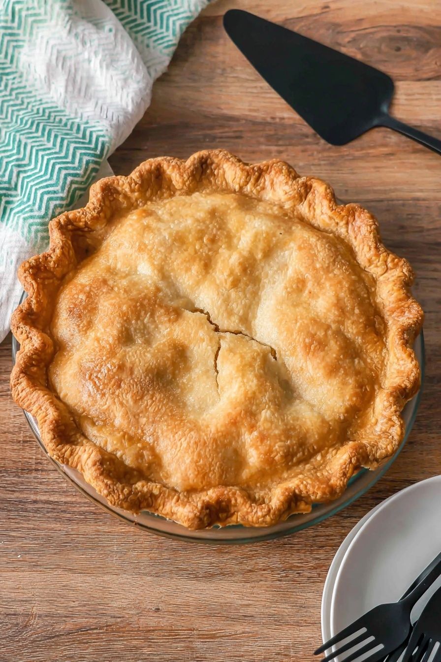 A whole golden-brown pie with a flaky, bubbly crust and a crimped edge sits centered on a clear glass pie dish. The crust has a slightly uneven, homemade texture with a few small cracks near the middle, showing a hint of the filling below. The pie is placed on a wooden surface, above which rests a black pie server with a matte finish. In the bottom right corner, there is a white plate holding three black forks, and to the left, a white cloth with a green chevron pattern peeks into the frame. photo taken with an iphone --ar 2:3 --v 7 - Pear Pie, Delicious Pear Pie, Fall Dessert Ideas, Easy Pear Pie Recipe, Cozy Fruit Pie