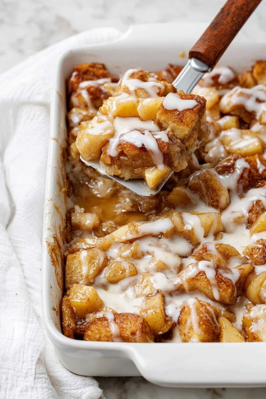 A white rectangular baking dish filled with a baked dessert made of soft, golden brown pieces layered with light brown cooked apple chunks and drizzled generously with a white creamy glaze. The dessert has a slightly sticky texture from syrup or sauce that pools around the pieces. A metal spatula with a wooden handle lifts a portion showing the soft inside layers and more apples. The background is a white marbled surface with a white cloth nearby. photo taken with an iphone --ar 2:3 --v 7 - Apple Pie Cinnamon Roll Bake, cinnamon roll breakfast bake, apple pie cinnamon roll, easy apple cinnamon breakfast, fall brunch recipes