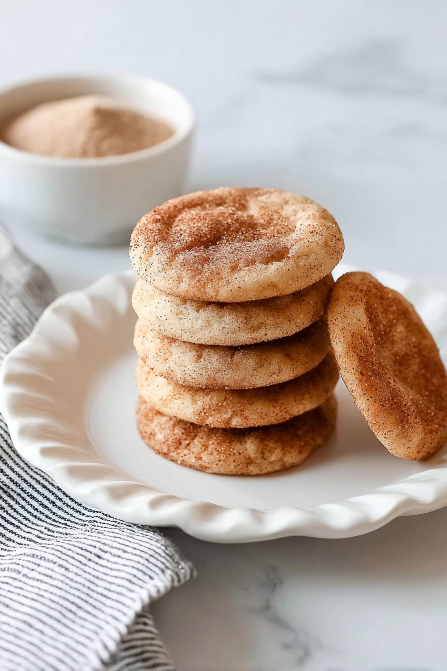 A stack of eight round, light brown cookies with a dusting of cinnamon sugar on top sits in the center of a white plate with scalloped edges. One cookie lies flat behind the stack, showing its darker cinnamon spots. The white plate is placed on a white marbled surface with a folded white and black striped cloth partially visible on the left. In the top left corner, a small white bowl filled with a light brown powder is slightly out of focus. photo taken with an iphone --ar 2:3 --v 7 - Chewy Snickerdoodle Cookies, cinnamon sugar cookies, soft chewy cookie recipe, homemade snickerdoodles, classic cinnamon cookies