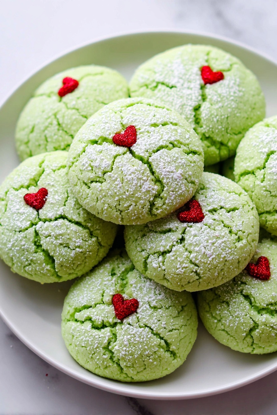 The image shows a white plate filled with round green cookies that have a cracked texture on top. Each cookie has a light dusting of white powder and a tiny red heart decoration placed slightly off-center on the top surface. The cookies are stacked closely together on the plate, which sits on a white marbled background. Photo taken with an iphone --ar 2:3 --v 7 - Grinch Peppermint Cookies, festive holiday cookies, green peppermint cookies, easy Christmas cookie recipe, colorful holiday treats