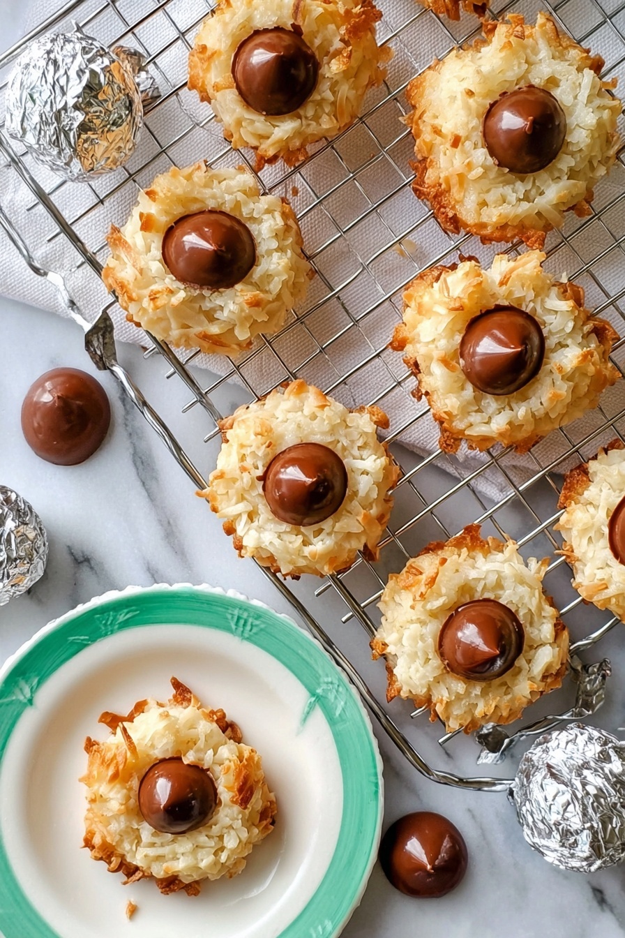 The image shows seven round coconut cookies with a light golden-brown crispy outside and white, shredded coconut texture in the middle layer. Each cookie has a single smooth milk chocolate drop placed in the center on top. The cookies are arranged on a silver cooling rack that rests on a white rectangular marble slab. Around the rack, there are several extra chocolate drops scattered on the white marbled surface. A woman's hand is holding one cookie on a white plate with a green rim, and a few silver-wrapped chocolate candies are placed nearby. The overall setting is bright with a white marbled background. photo taken with an iphone --ar 2:3 --v 7 - Coconut Macaroon Blossoms with Chocolate Kisses, coconut macaroon cookies, chocolate kiss cookies, gluten free coconut cookies, easy holiday cookie recipes