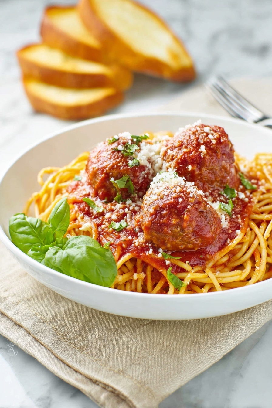 A white bowl holds a bed of golden-yellow spaghetti noodles topped with rich red tomato sauce. On top, there are three large brown meatballs coated in the sauce, sprinkled with white grated cheese and small green herb pieces. A fresh green basil leaf adds color near the front. In the background, there are three toasted bread slices leaning against the bowl. The scene sits on a beige cloth over a white marbled surface. A silver fork rests nearby on the right side. Photo taken with an iphone --ar 2:3 --v 7 - Homemade Spaghetti and Meatballs, Italian pasta with meatballs, easy spaghetti and meatballs, classic spaghetti and meatballs, hearty Italian dinner