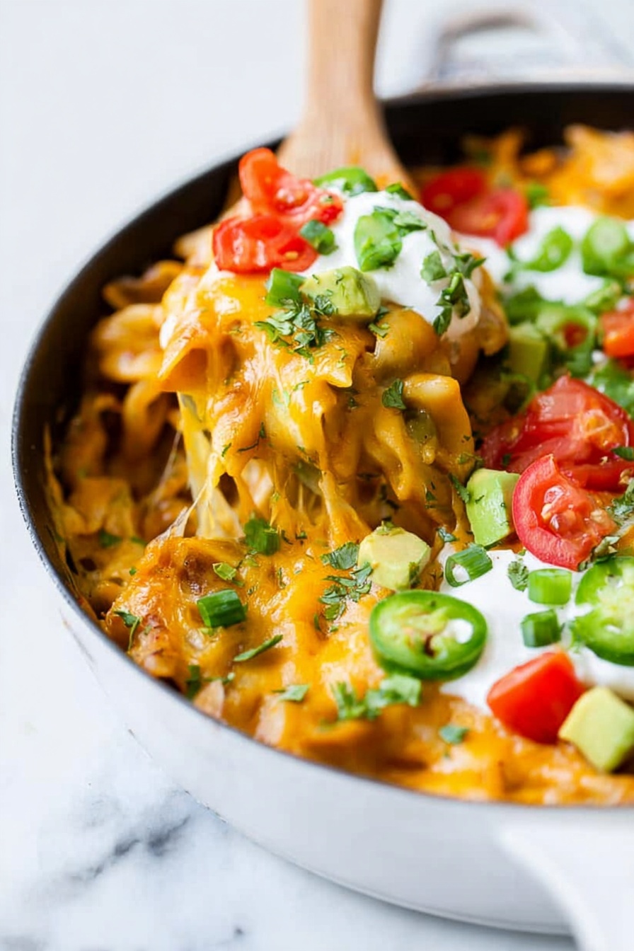 A white pan filled with baked pasta covered in melted orange cheese that stretches as a wooden spatula lifts a serving. On top, there are chopped green herbs and sliced green onions, bright red chopped tomatoes, creamy white sour cream, and small chunks of green avocado layered over the cheesy pasta. The background is a white marbled surface. photo taken with an iphone --ar 2:3 --v 7 - Cheesy Enchilada Pasta Skillet, cheesy enchilada pasta, easy enchilada pasta dinner, quick pasta skillet, one-pot enchilada pasta