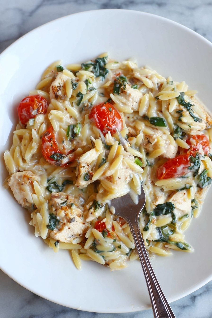 A white round plate holds a creamy pasta dish composed of orzo pasta mixed with small chunks of light brown cooked chicken pieces and bright red cherry tomatoes. There are dark green leafy bits, possibly spinach or herbs, spread evenly throughout the dish. The sauce looks smooth and creamy, coating the pasta and ingredients in a soft white layer. A silver fork is placed inside the pasta near the bottom center of the plate. The plate sits on a white marbled surface photo taken with an iphone --ar 2:3 --v 7 - Creamy Chicken Orzo Bake, Chicken Orzo casserole, cheesy chicken bake, easy weeknight chicken dinner, comforting baked chicken dish