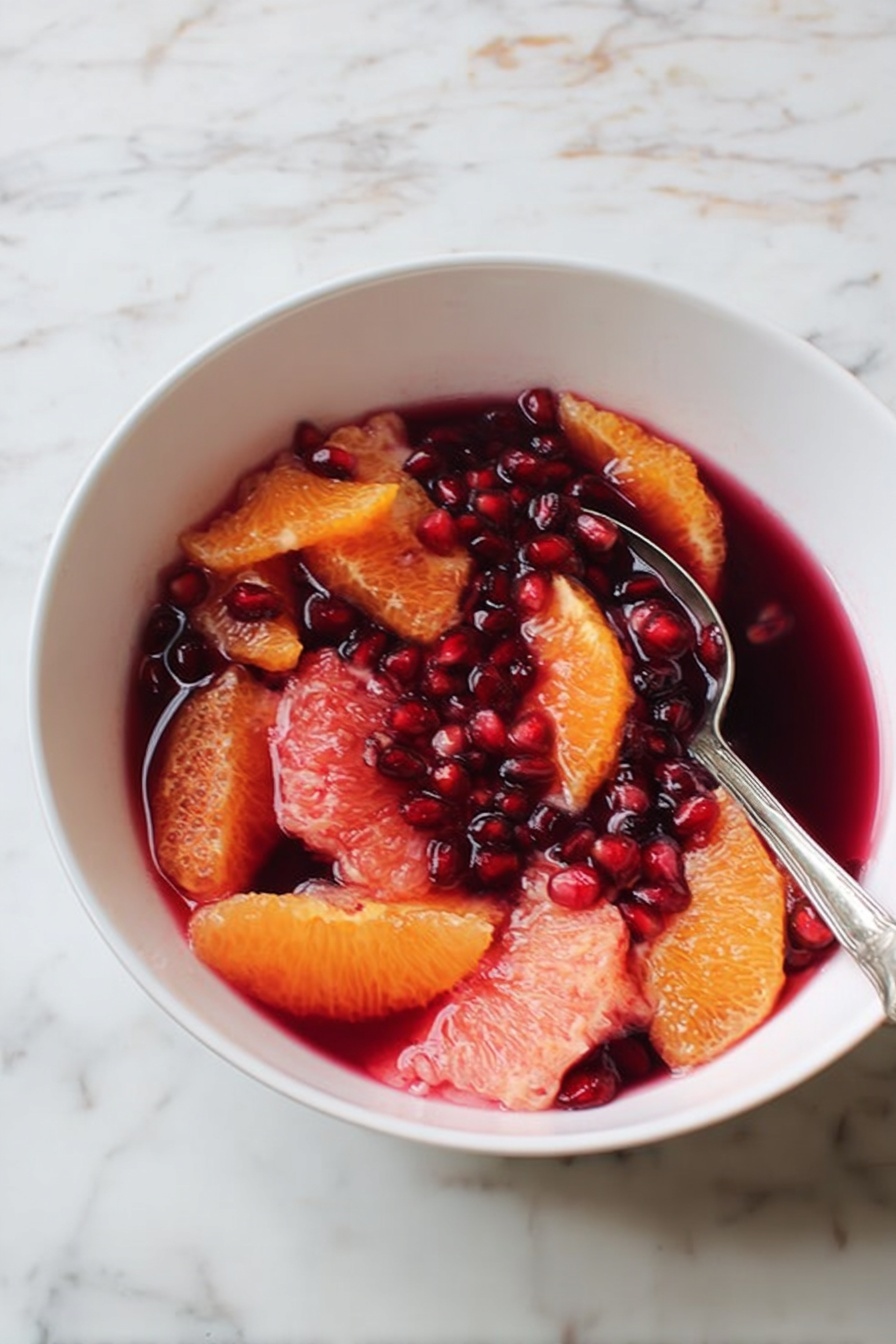 A white bowl filled with two main layers: the bottom layer is a deep red liquid, and floating on top are bright orange and pink citrus slices along with many shiny dark red pomegranate seeds. A silver spoon rests inside the bowl on the right side. The bowl is placed on a white marbled surface with soft veins of gray and faint brown. photo taken with an iphone --ar 2:3 --v 7 - Winter Citrus and Pomegranate Salad, citrus winter salad, pomegranate fruit salad, healthy winter salads, festive citrus salad