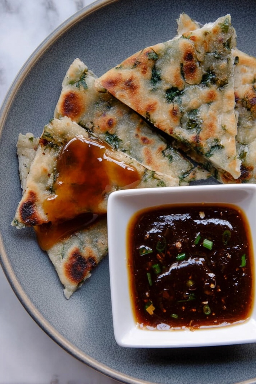 The image shows a close-up of a grey plate on a white marbled surface, with four triangular pieces of flatbread that have green herbs inside, each piece varying in size and texture with some browned spots from cooking. One piece near the bottom left has a shiny layer of sticky, dark amber sauce on top. To the right, a small white square bowl holds a thick, dark brown dipping sauce with small green bits inside, creating a glossy surface. photo taken with an iphone --ar 2:3 --v 7 - Chinese Scallion Pancakes, scallion pancake recipe, homemade scallion pancakes, crispy scallion pancakes, savory scallion pancakes